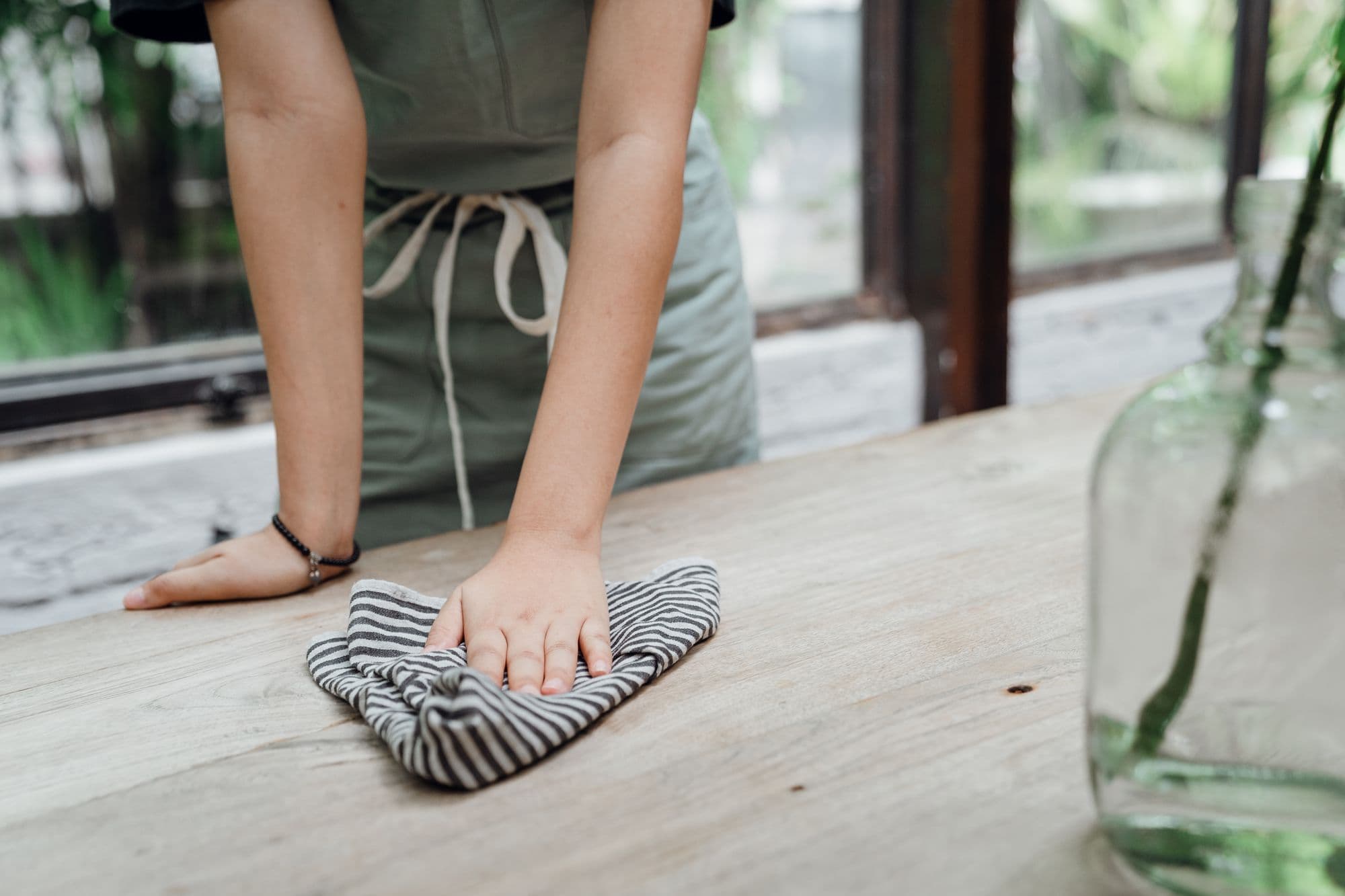 Gros plan sur les mains d’une personne en tablier vert-gris qui essuie une table en bois brut avec un torchon rayé ; le visage est hors cadre, un vase en verre apparaît au premier plan à droite.