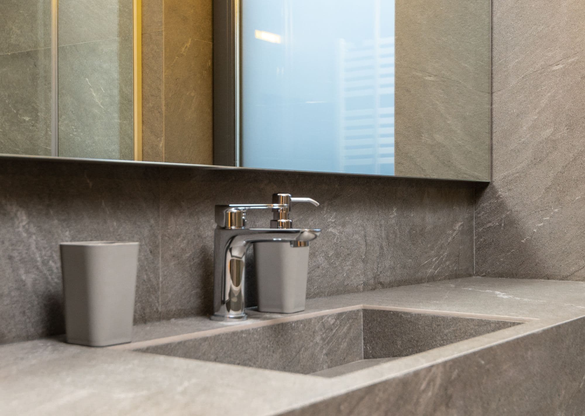 Détail de salle de bains : vasque rectangulaire taillée dans un plan en pierre grise, mitigeur chromé, deux gobelets gris et mur en carreaux de pierre sous un miroir.
